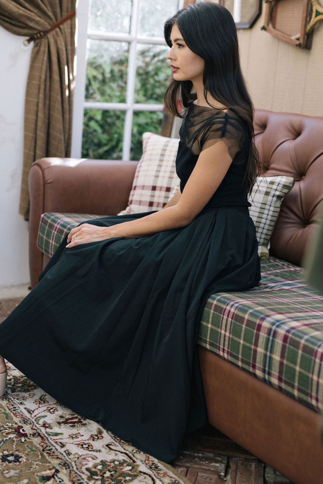 The image shows a woman wearing a long, flowing black dress with sheer mesh detailing on the shoulders and sleeves. She is seated on a plaid sofa against a window, creating a cozy, intimate atmosphere.
