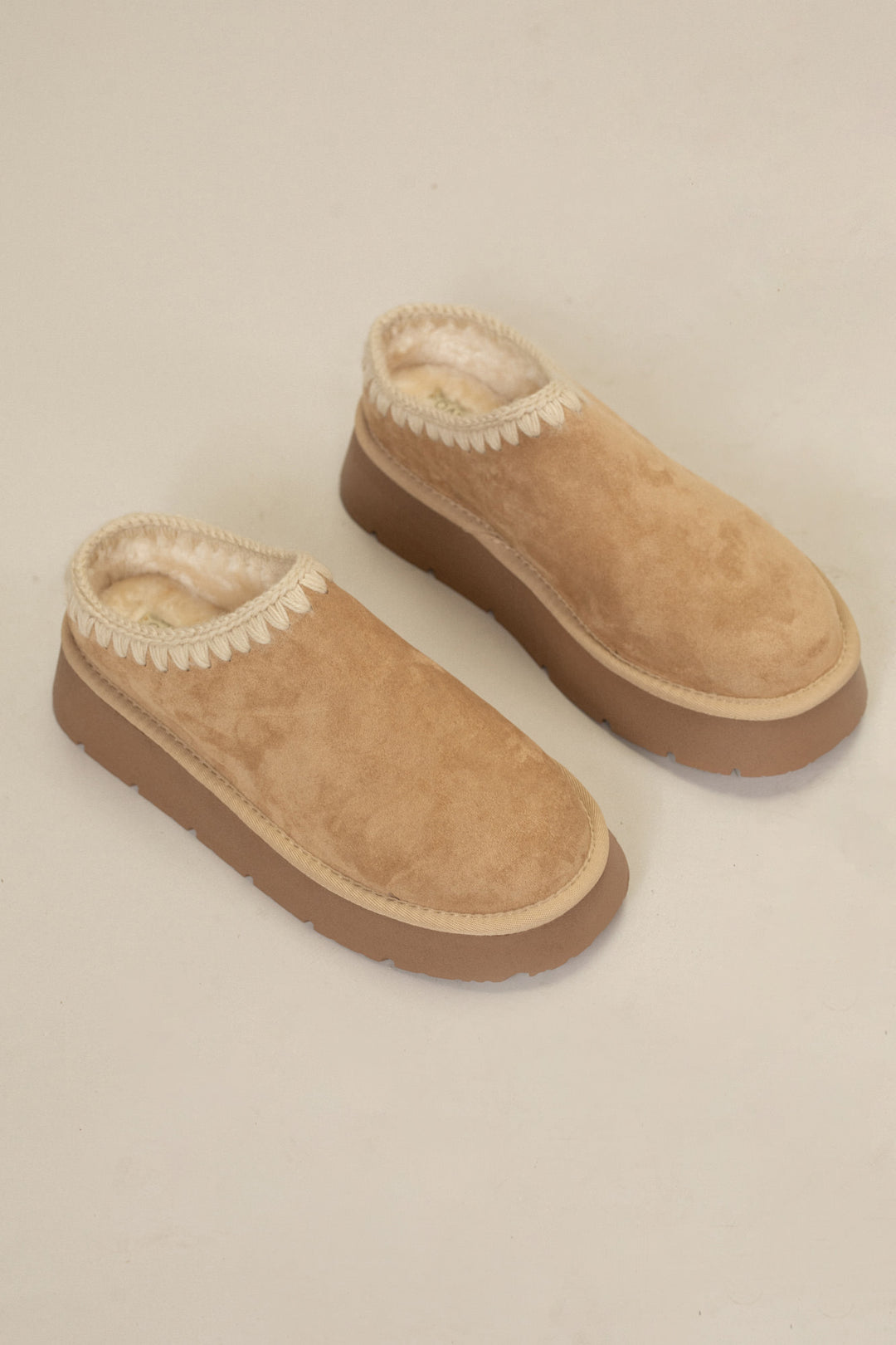 Tan suede platform mule slippers with cozy wool lining and decorative stitching around the collar.