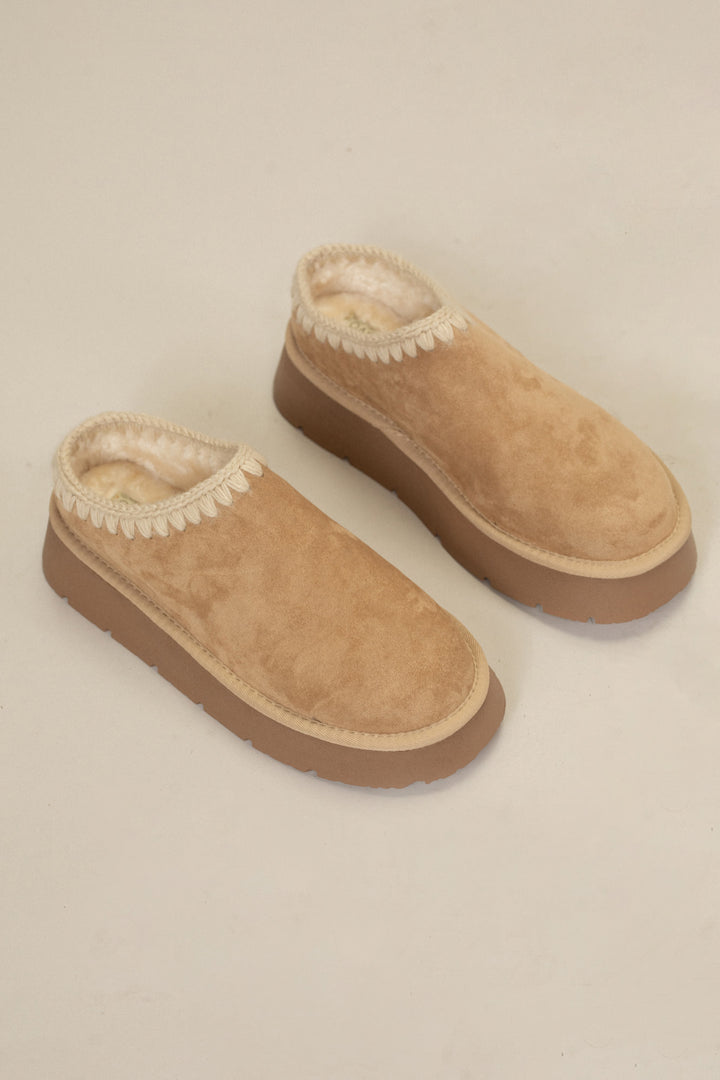 Tan suede platform mule slippers with cozy wool lining and decorative stitching around the collar.