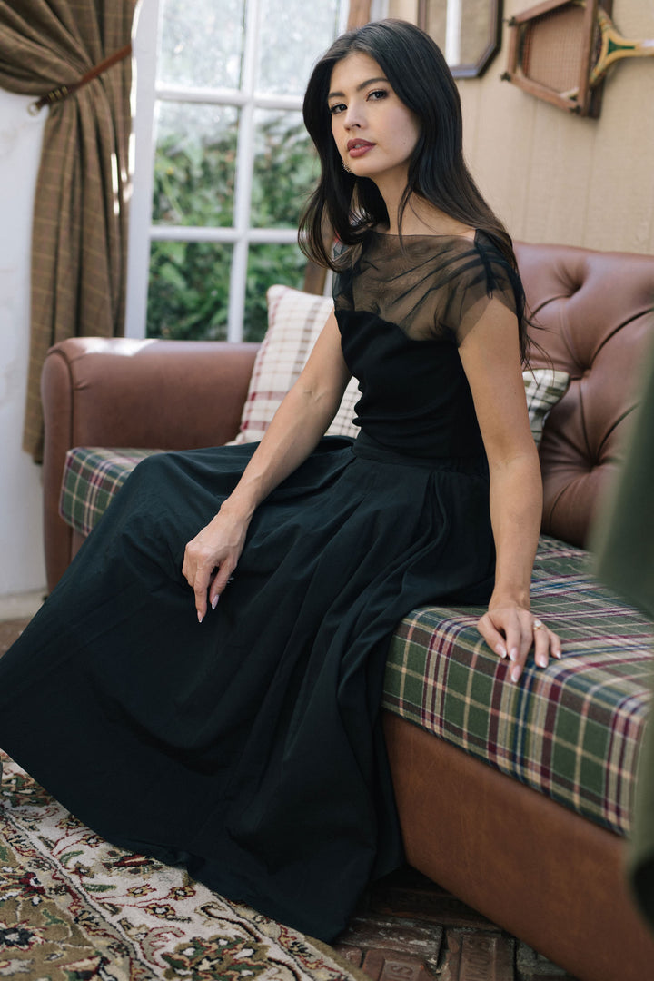 A woman wearing a black dress with a sheer, mesh-like top and long, flowing skirt sitting on a patterned couch against a window.