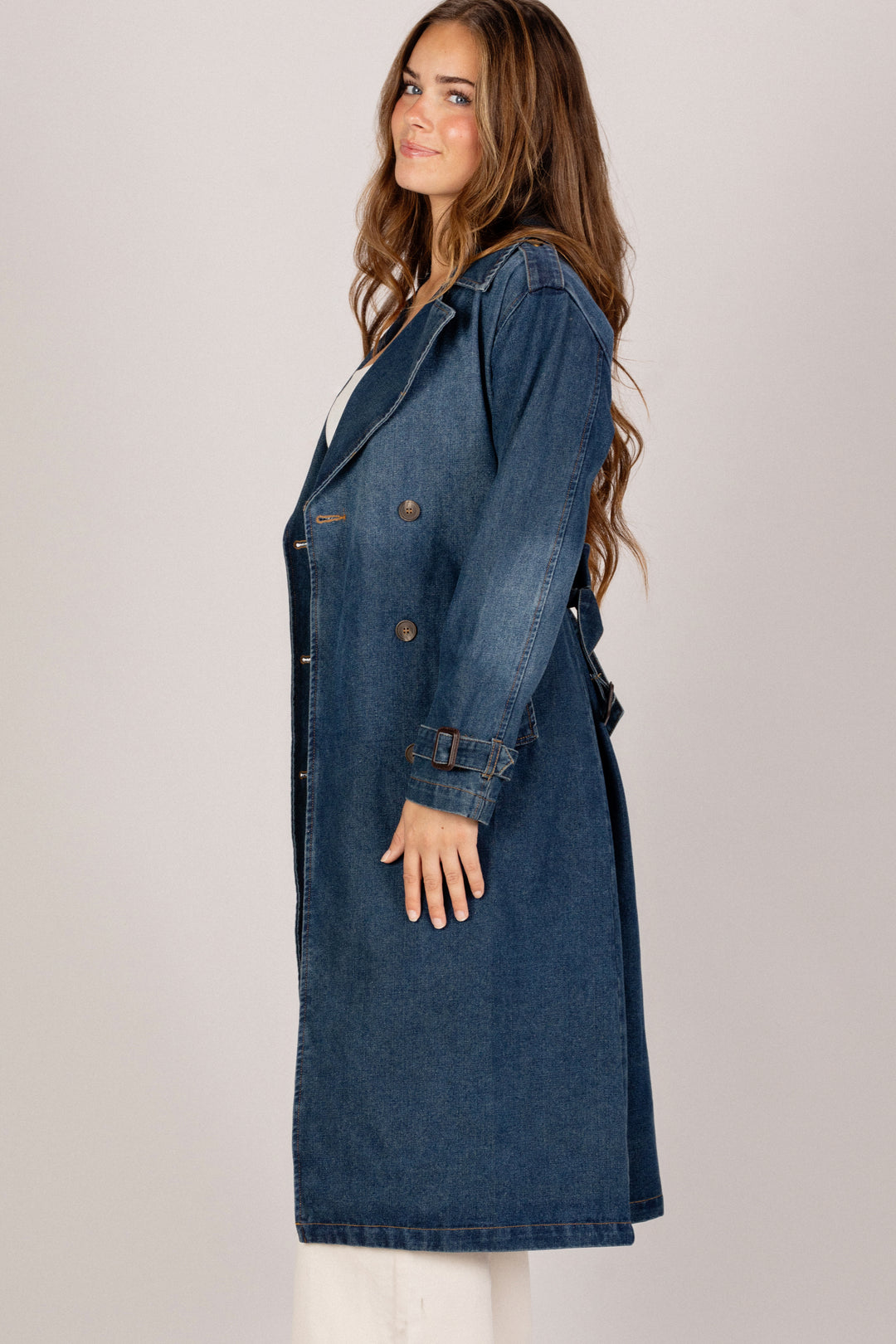 A young woman wearing a long, blue denim trench coat with a belted waist and button details.