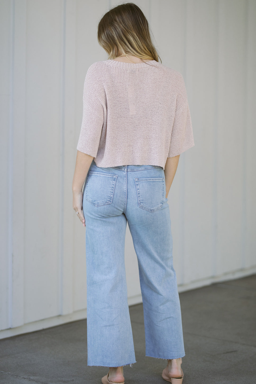 The image shows a woman wearing a light pink cropped cardigan with ribbed detailing and a V-neck. She is also wearing light wash high-rise jeans with a wide leg silhouette.