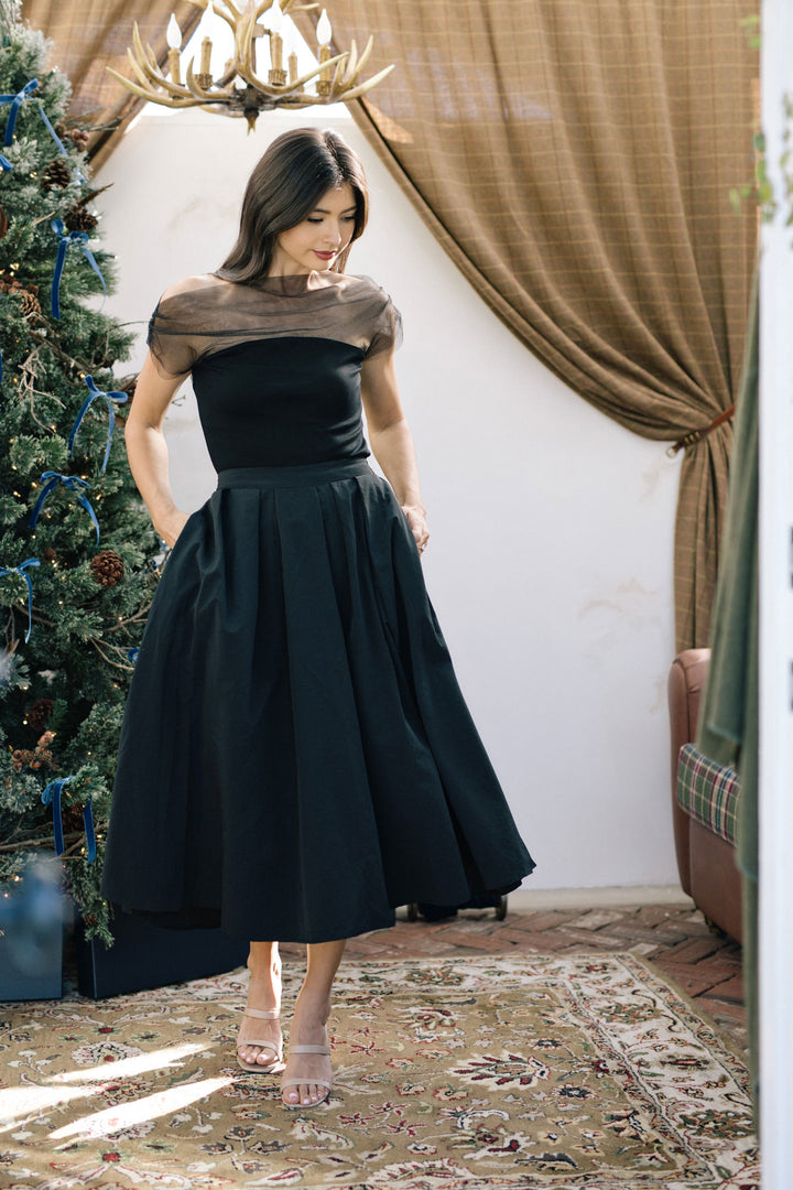 A woman in a black off-the-shoulder dress stands in front of a Christmas-decorated backdrop, wearing sandals and with her long dark hair down.