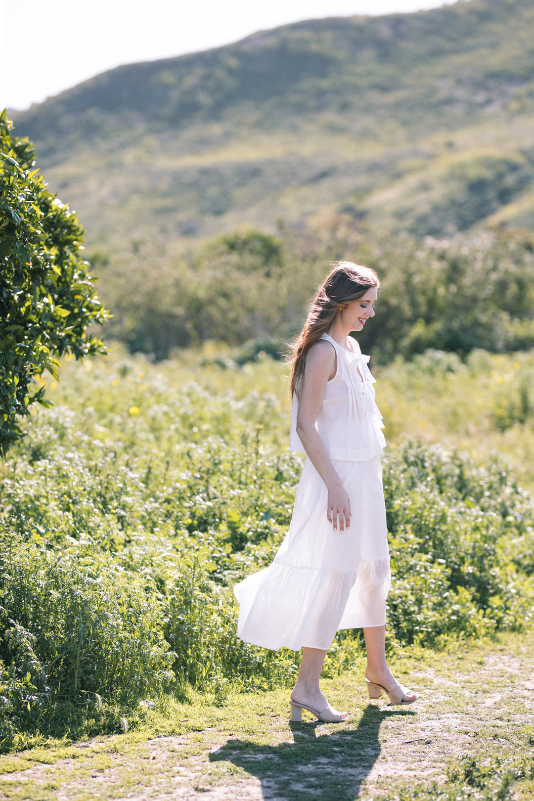 A young woman in a white, flowing tiered midi dress stands in a lush, green meadow, with mountains in the background.