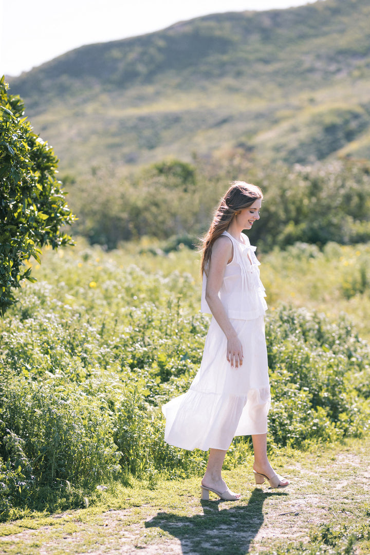 A young woman in a white, flowing tiered midi dress stands in a lush, green meadow, with mountains in the background.