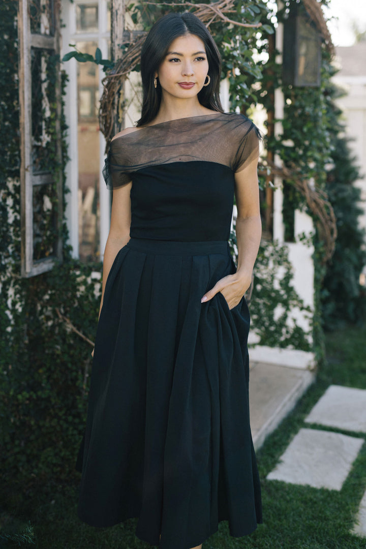 A woman in a long black dress with a sheer mesh neckline, standing among lush greenery.