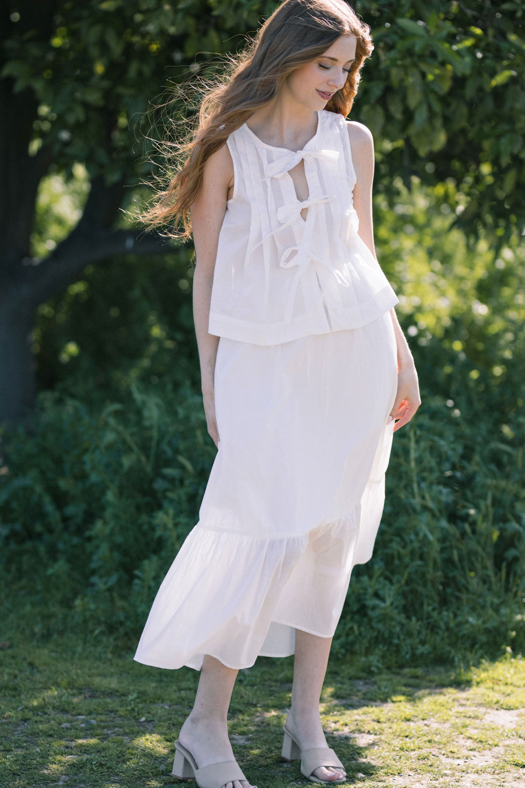 A young woman wearing a flowing, white sleeveless dress with ruffled details, standing in a lush green garden.
