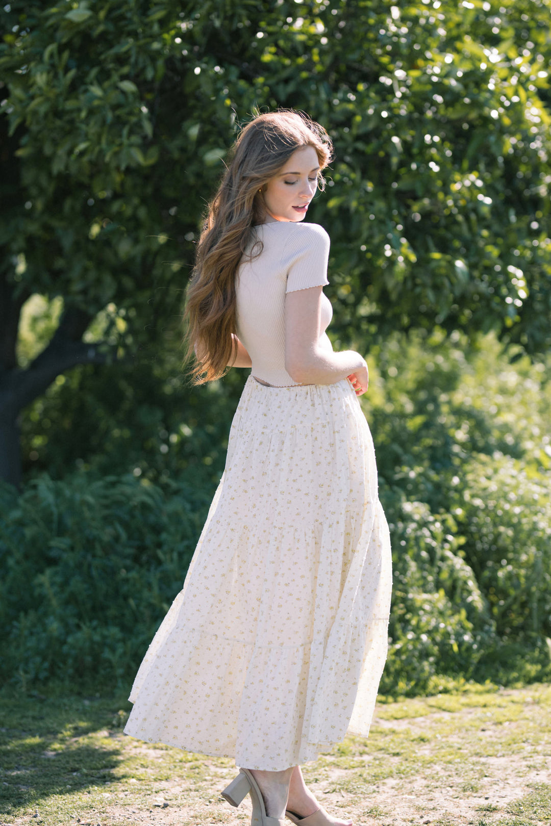 A woman with long hair stands in a lush green setting, wearing a cream-colored floral midi skirt and a fitted white top. The image showcases the details and flow of the tiered skirt design.