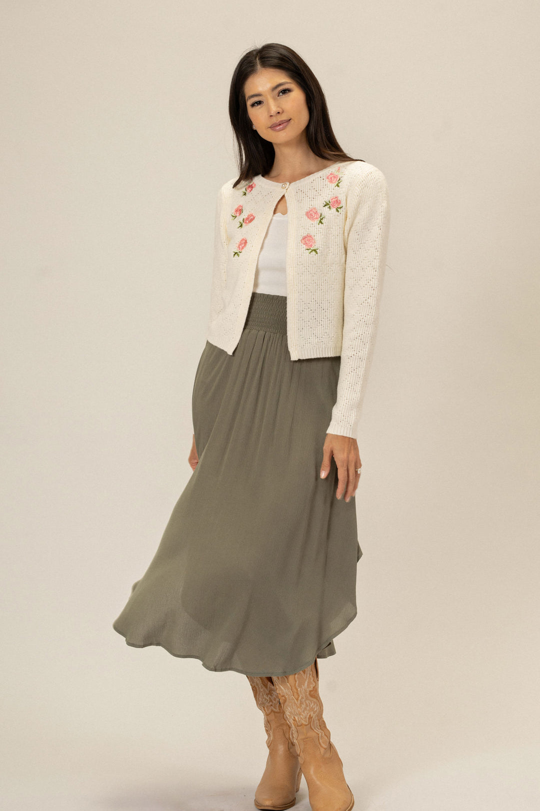 A white knit cardigan with pink floral embroidery, worn over a gray maxi skirt and paired with tan lace-up boots by a young woman with long dark hair.