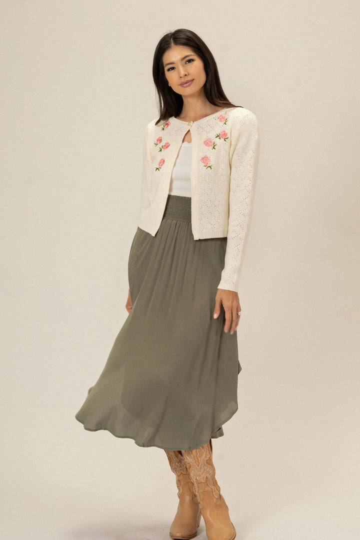 A white knit cardigan with pink floral embroidery, worn over a gray maxi skirt and paired with tan lace-up boots by a young woman with long dark hair.