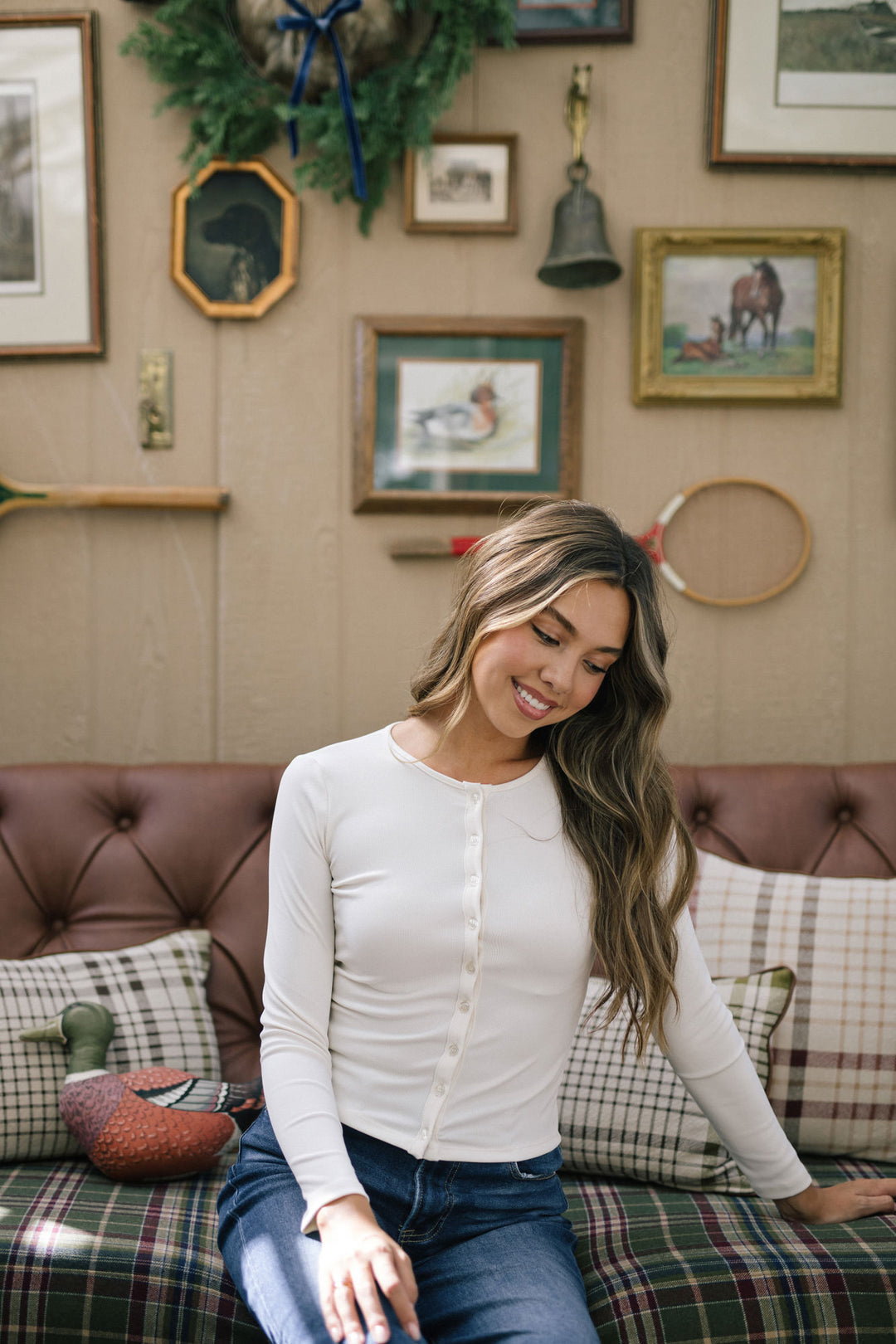 A smiling young woman with long wavy hair models a white ribbed button-up top in a cozy living room setting.