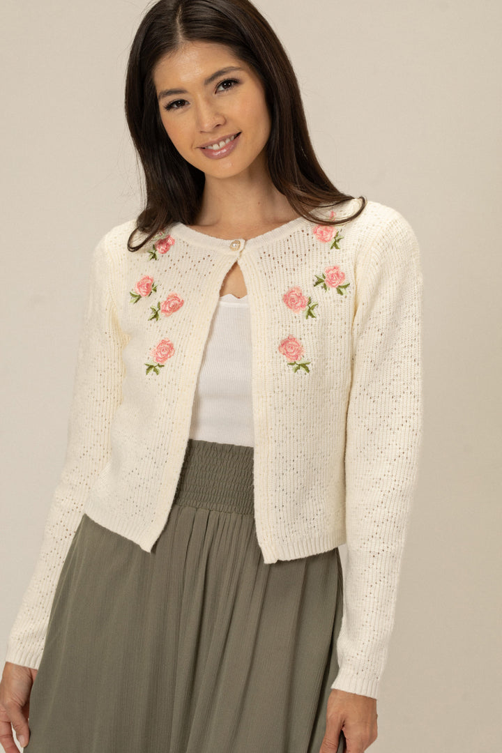 A white knit cardigan with pink floral embroidery, featuring a keyhole neckline, button closure, and fitted, cropped silhouette.