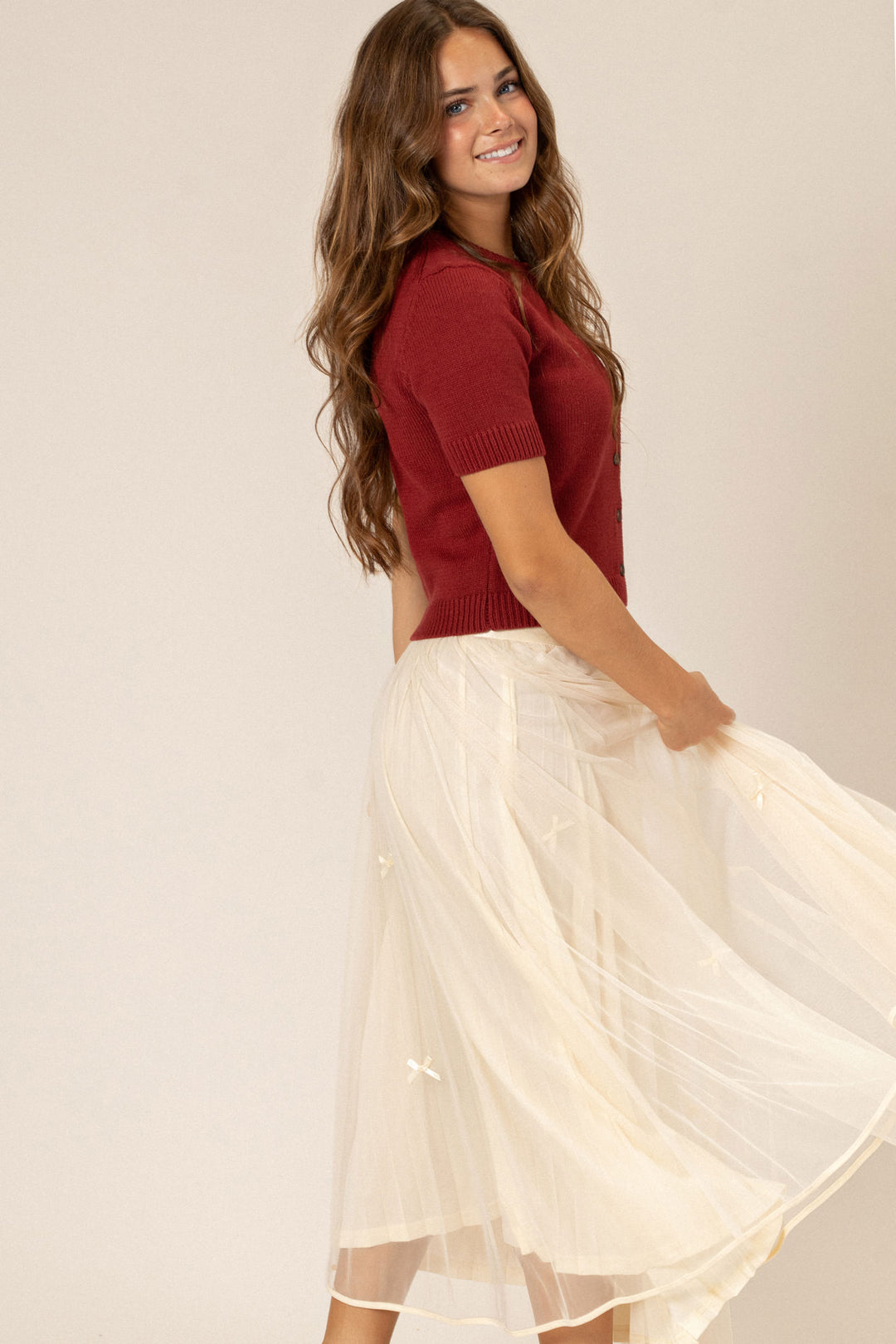 A young woman with long, wavy brown hair smiles while wearing a red knit top with a button neckline and a mid-length, flowy white skirt.