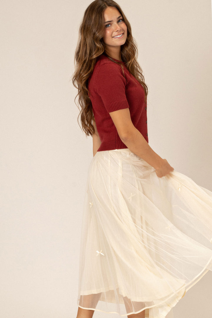 A young woman with long, wavy brown hair smiles while wearing a red knit top with a button neckline and a mid-length, flowy white skirt.