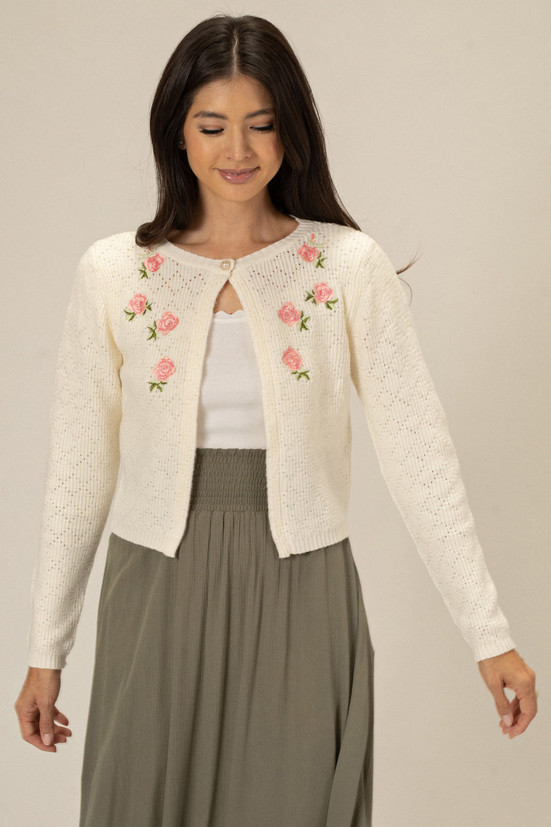 A white knitted cardigan with delicate pink rose embroidery, worn by a woman with long dark hair against a neutral background.