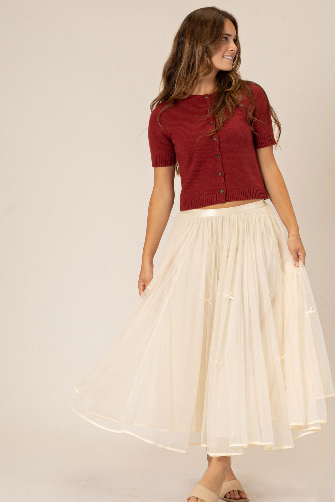 A young woman in a red knit cardigan top and an elegant cream-colored tulle skirt, smiling and posing confidently.
