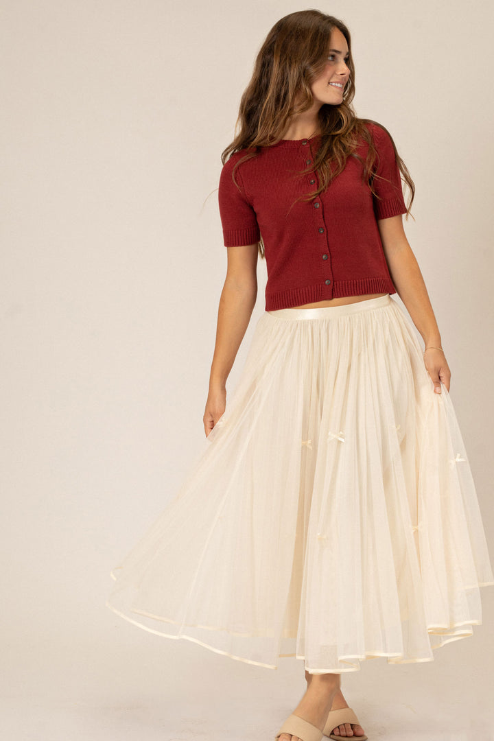 A young woman in a red knit cardigan top and an elegant cream-colored tulle skirt, smiling and posing confidently.