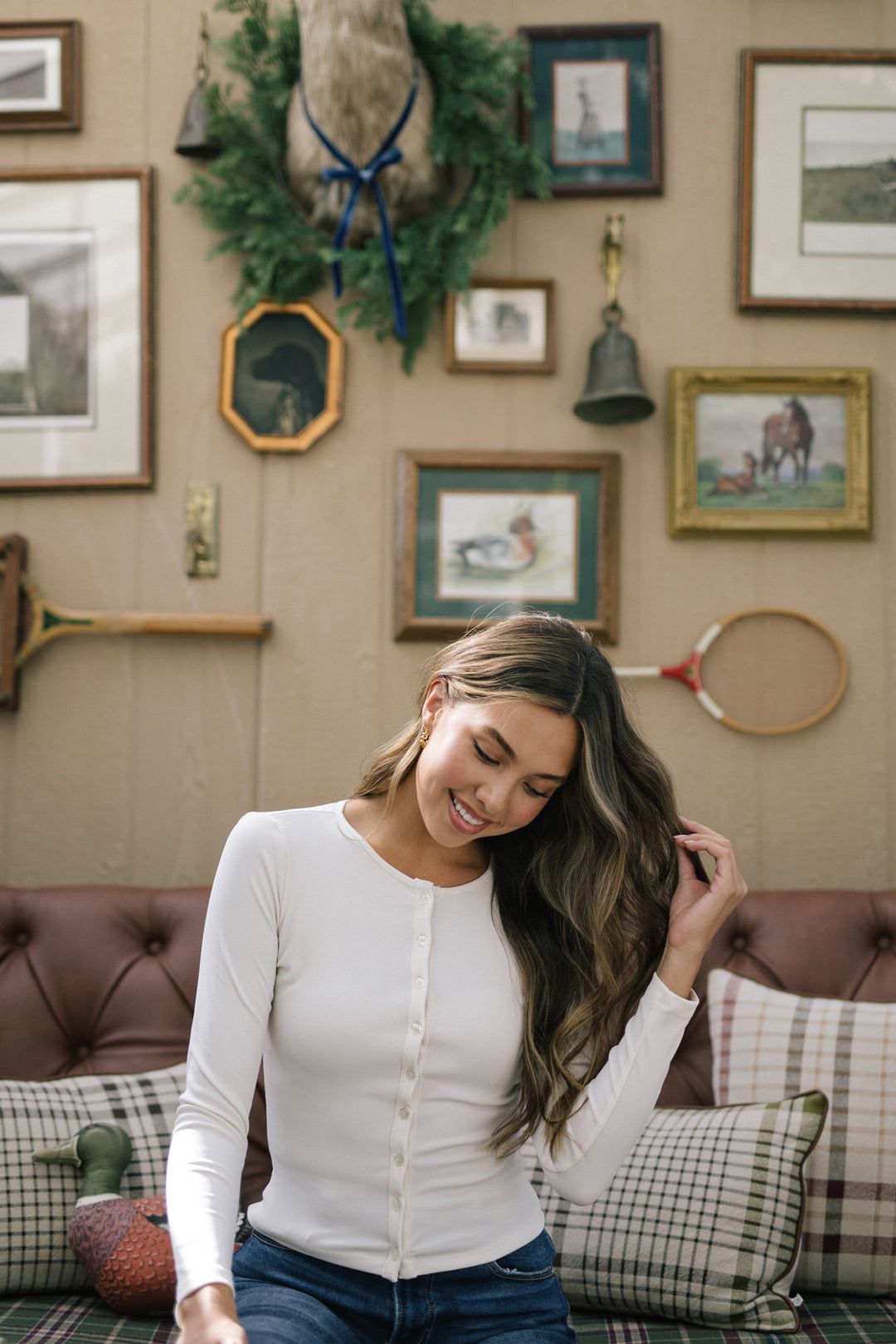 A smiling young woman with long, wavy brown hair models a white ribbed button-up top in a cozy, gallery-inspired living room setting.