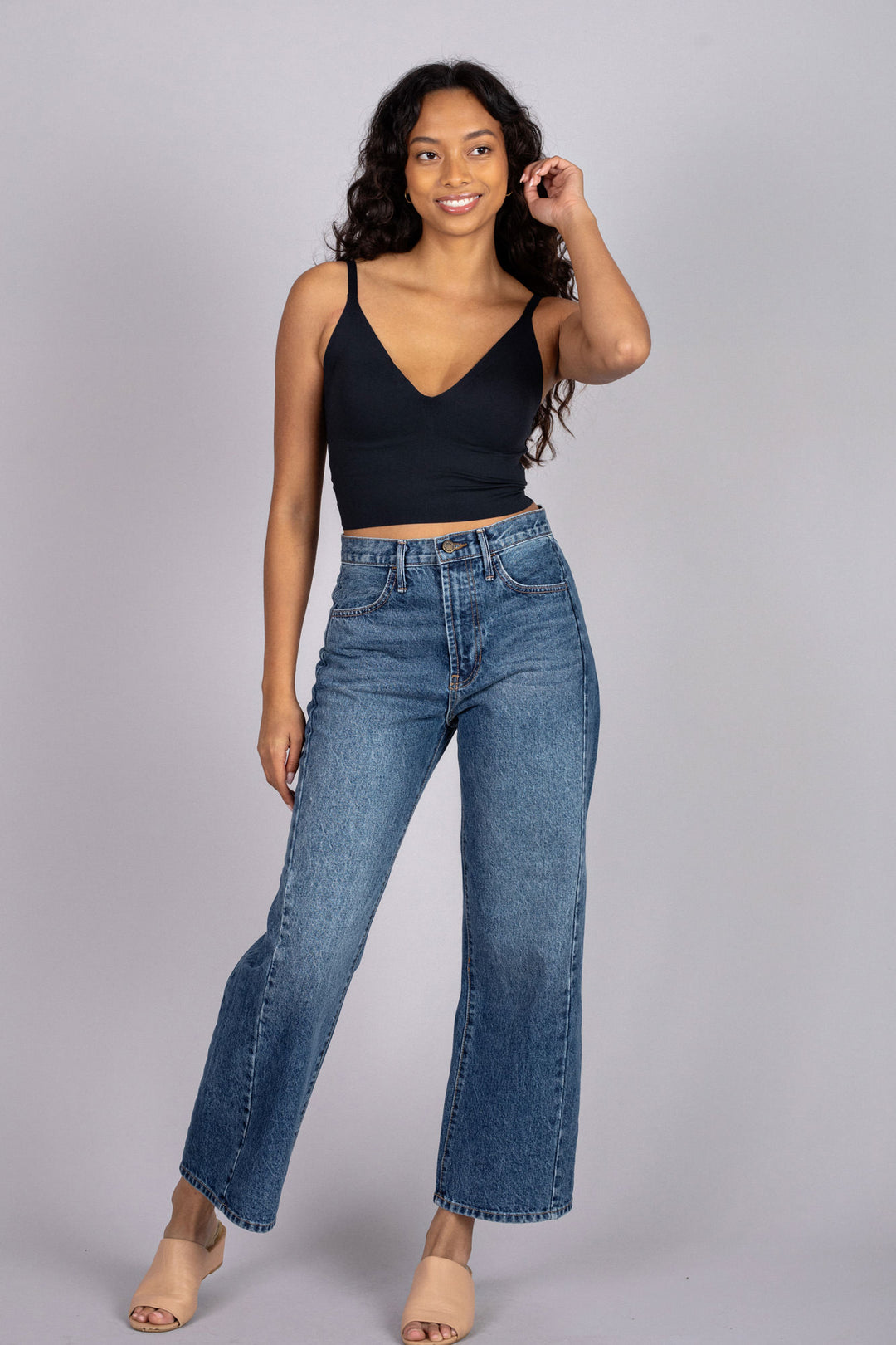 A young woman with long, dark curly hair wears a black tank top and high-waisted blue jeans, smiling at the camera.