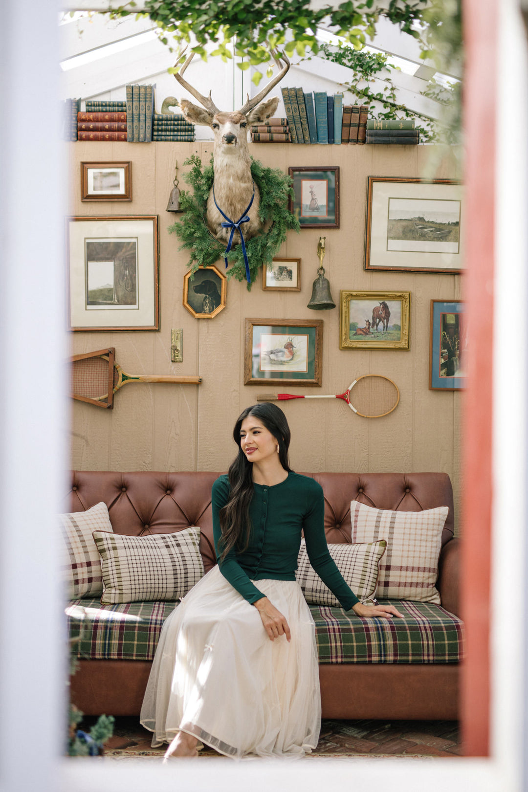 A young woman in a green long-sleeved top and a white midi skirt sits on a plaid sofa in a cozy room decorated with antiques and artwork.