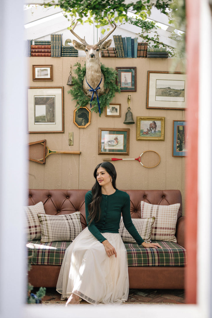 A young woman in a green long-sleeved top and a white midi skirt sits on a plaid sofa in a cozy room decorated with antiques and artwork.