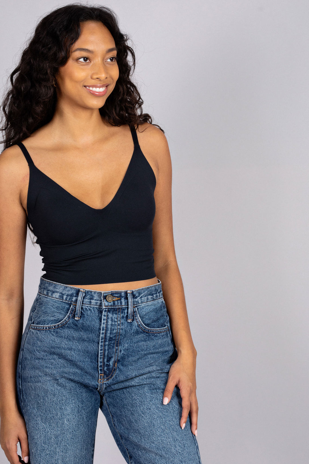 A young woman wearing a black sleeveless tank top and high-waisted blue jeans, smiling confidently against a gray background.