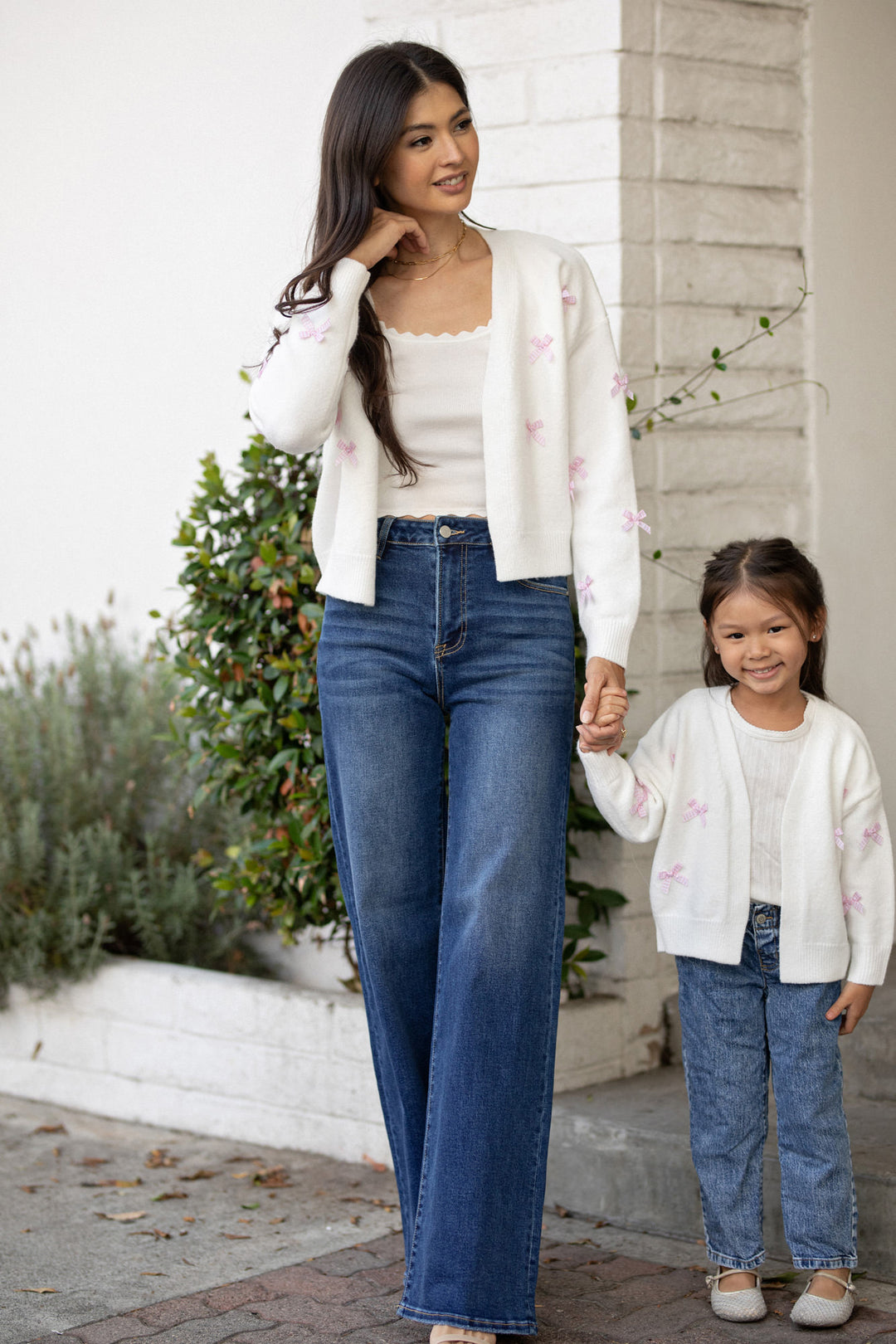 A woman in a white cardigan with pink floral embroidery holds hands with a young girl in a matching cardigan, both smiling against a backdrop of greenery.