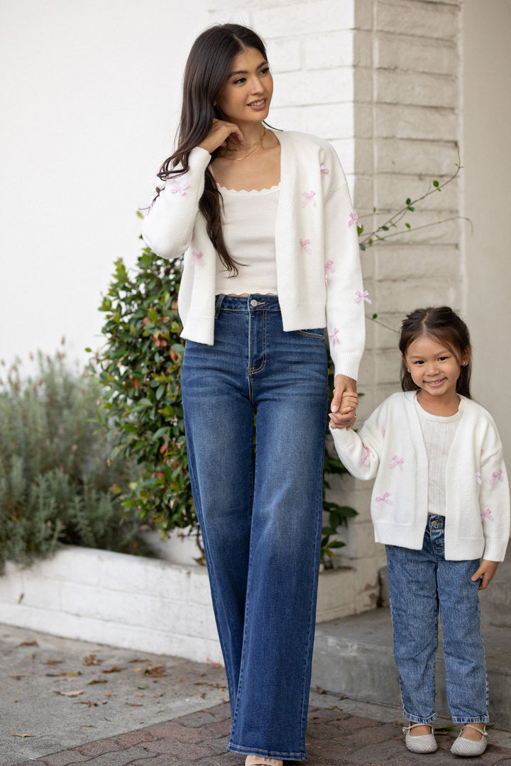 A woman in a white cardigan with pink floral embroidery holds hands with a young girl in a matching cardigan, both smiling against a backdrop of greenery.