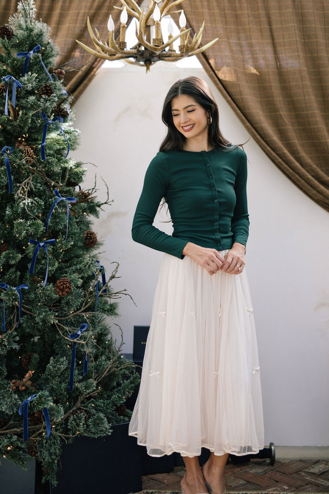 A woman wearing a green, long-sleeved button-up top and a full white tulle skirt, standing in front of a Christmas tree with twinkling lights.