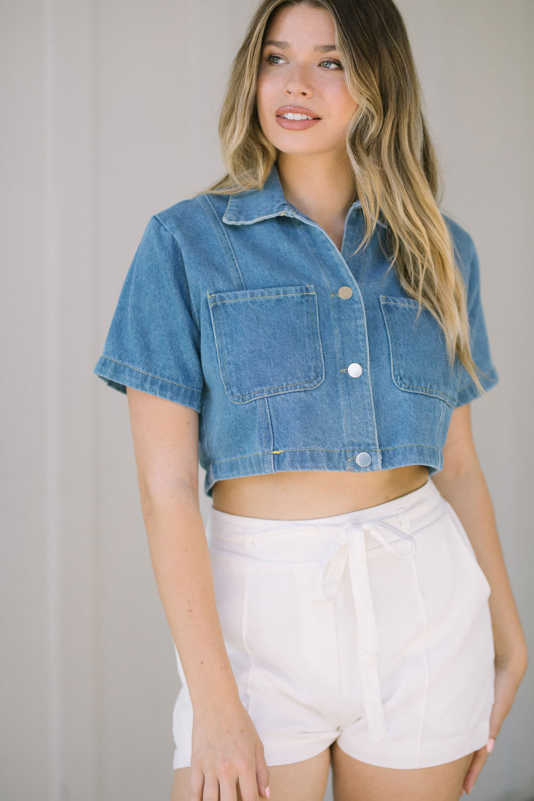 A young woman wearing a cropped denim shirt with short sleeves, paired with white high-waisted shorts, stands against a light background.