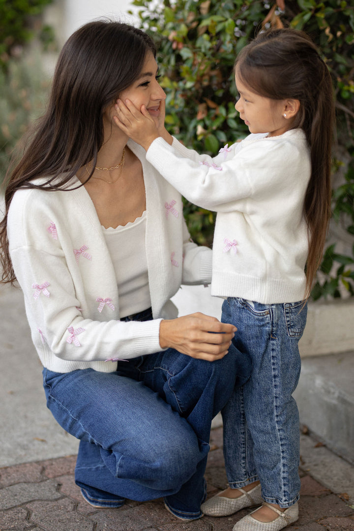 A young girl in a white cardigan with pink bows smiles as she gently touches the face of another young girl wearing a white sweater.
