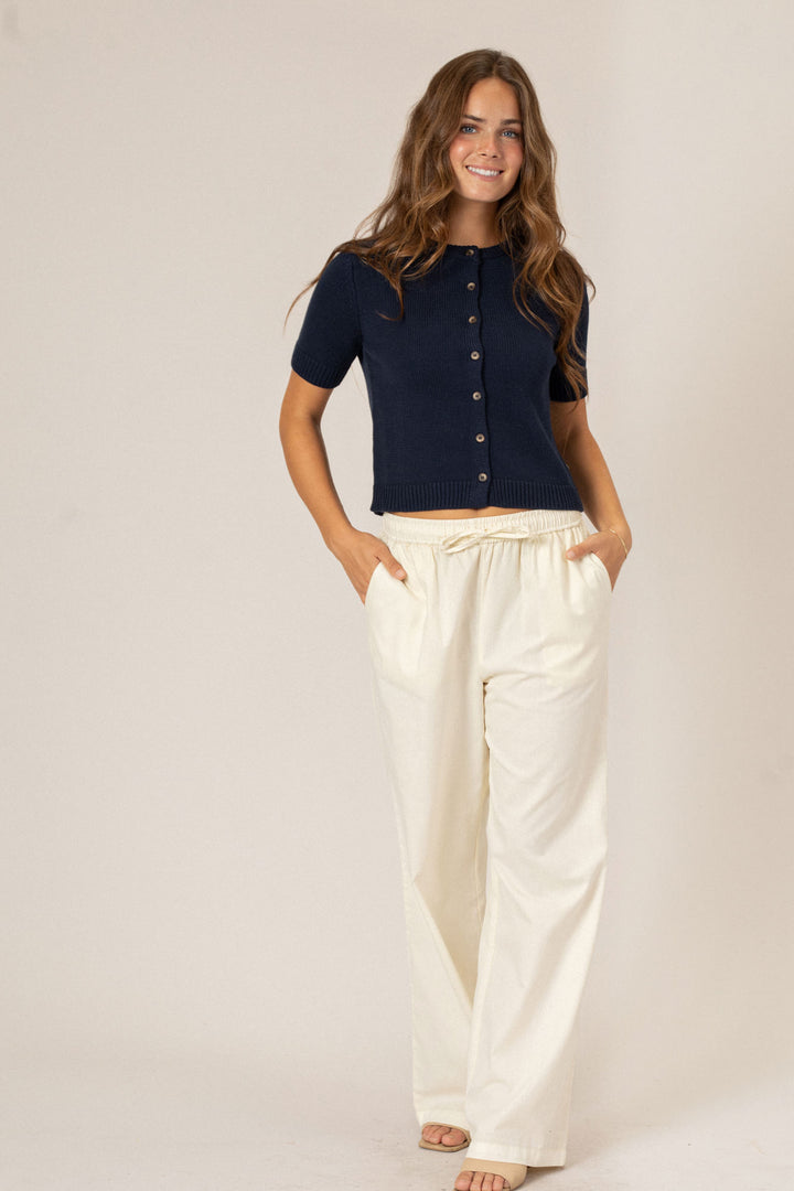 A smiling young woman wearing a navy blue buttoned knit top and off-white wide-leg pants, standing with her hands in the pockets.