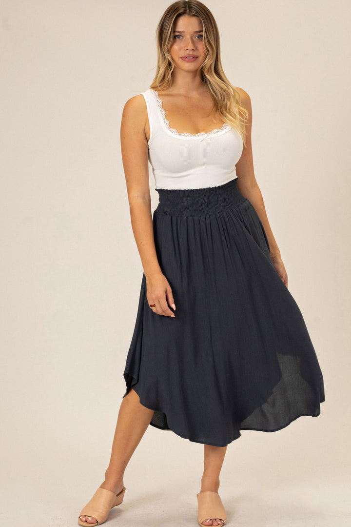 A young woman with long, wavy blonde hair wearing a white tank top and a flowing, dark blue midi skirt with an elastic waistband and a round hemline.
