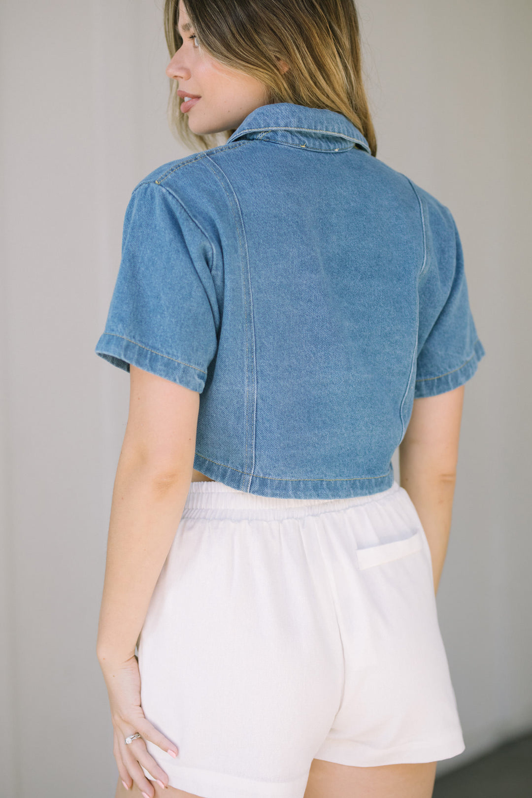 A cropped denim shirt with a collared neckline and short sleeves, worn with a pair of white shorts.