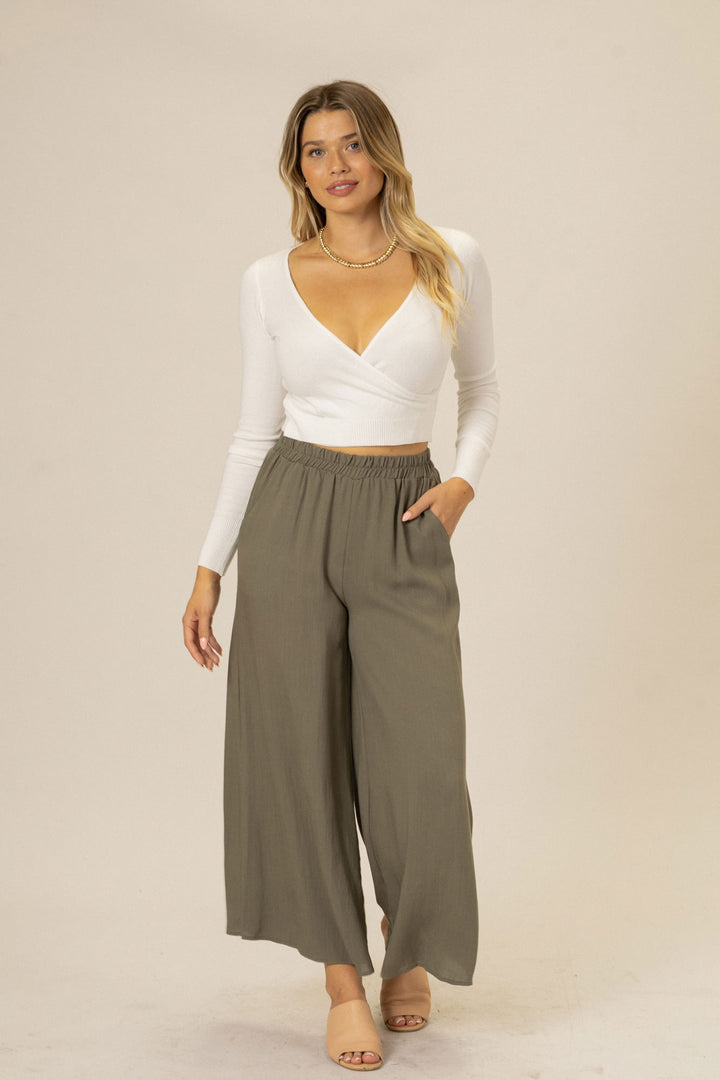 A woman wearing a white cropped top and gray wide-leg pants, standing with her hands in the pockets.