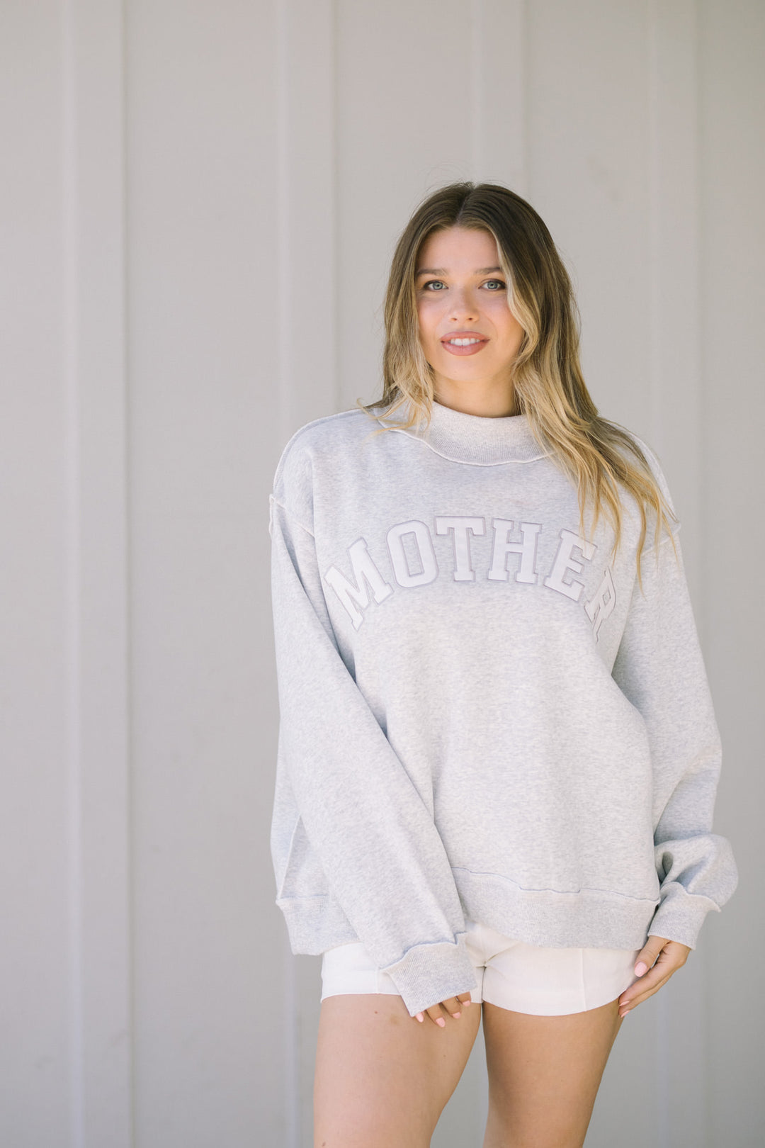 Woman wearing a gray sweatshirt with "MOTHER" text printed in white, standing against a plain background.