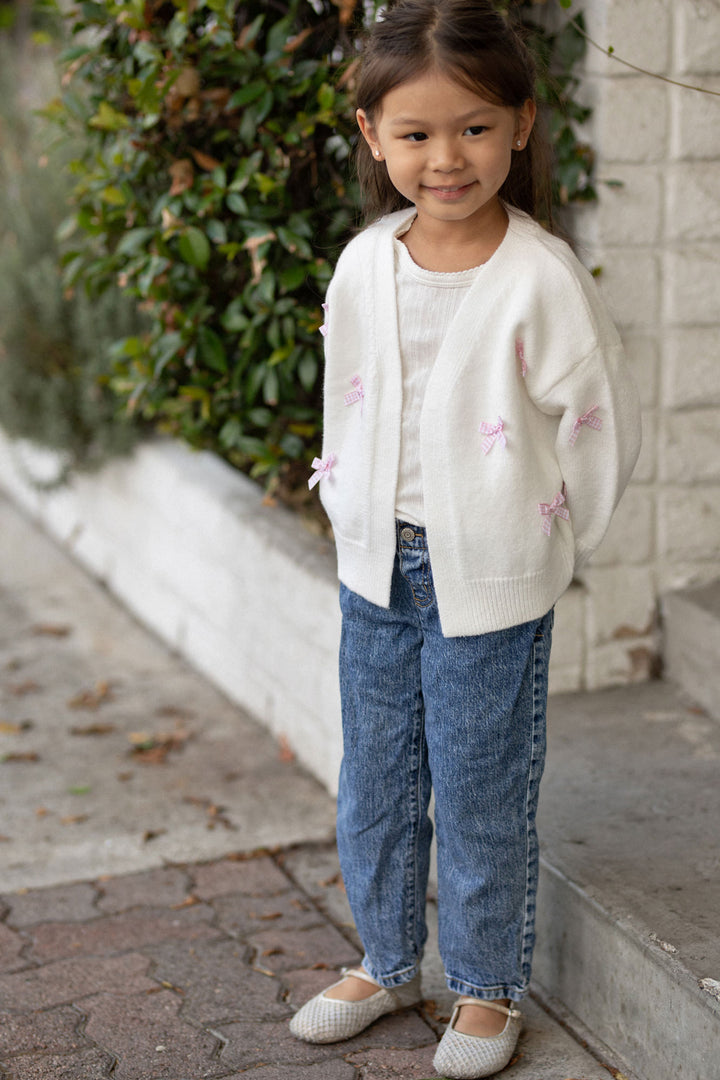 A young girl stands smiling, wearing a soft white cardigan with pink bows, paired with blue jeans and white shoes.