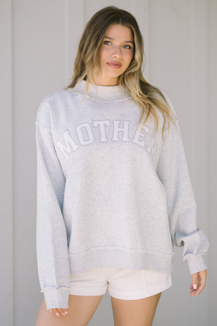 Gray mock-neck sweatshirt with "MOTHER" text, modeled by a woman with long blonde hair.