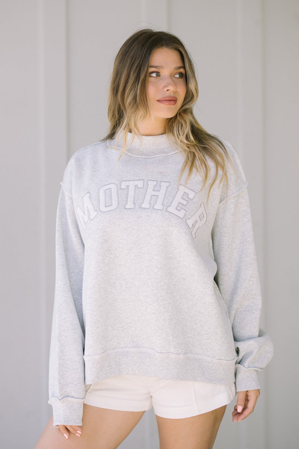 A gray mock-neck sweatshirt with "MOTHER" printed in white lettering, worn by a young woman with long blonde hair.