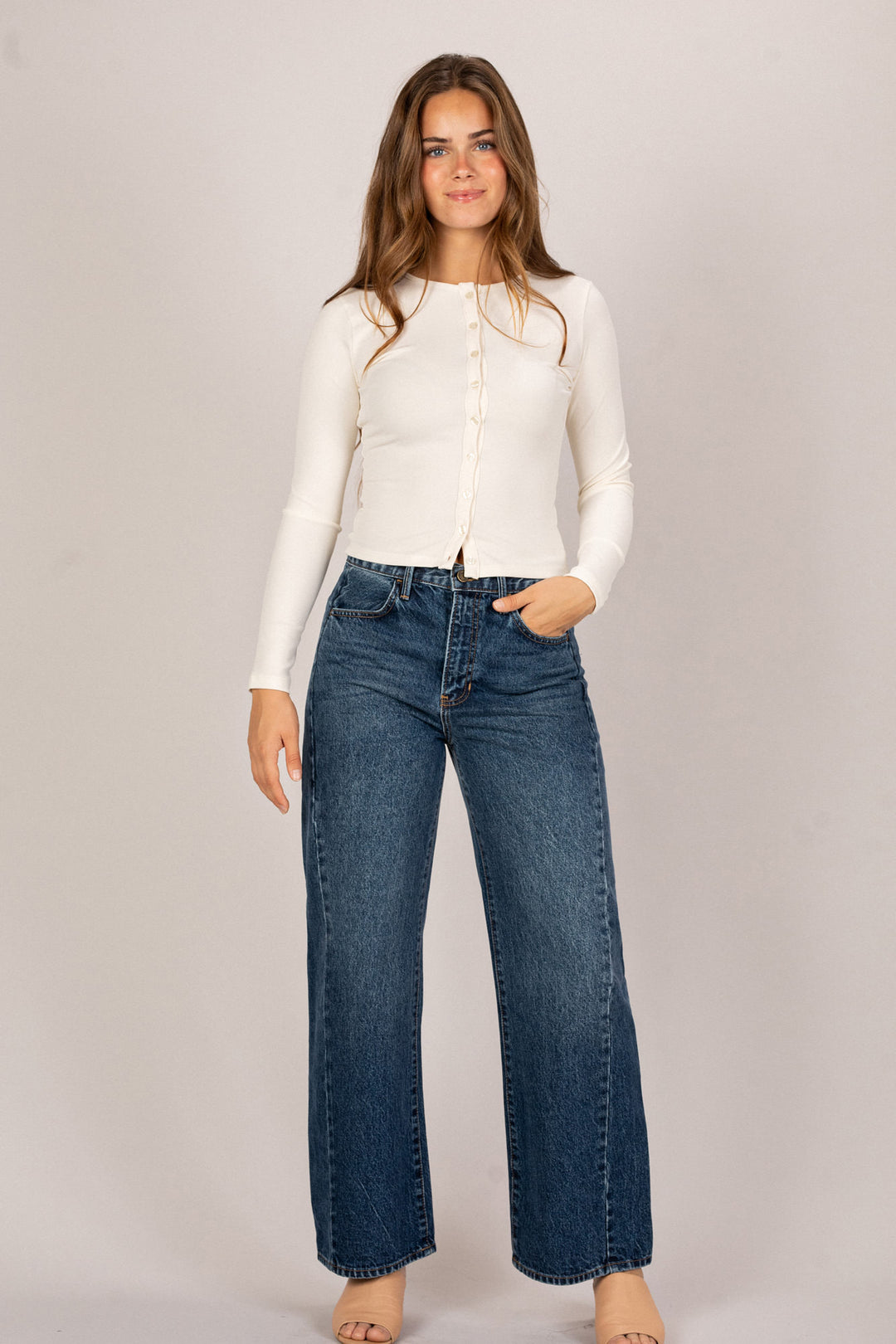 A young woman wearing a white long-sleeved shirt and dark denim wide-leg jeans, standing confidently against a neutral background.