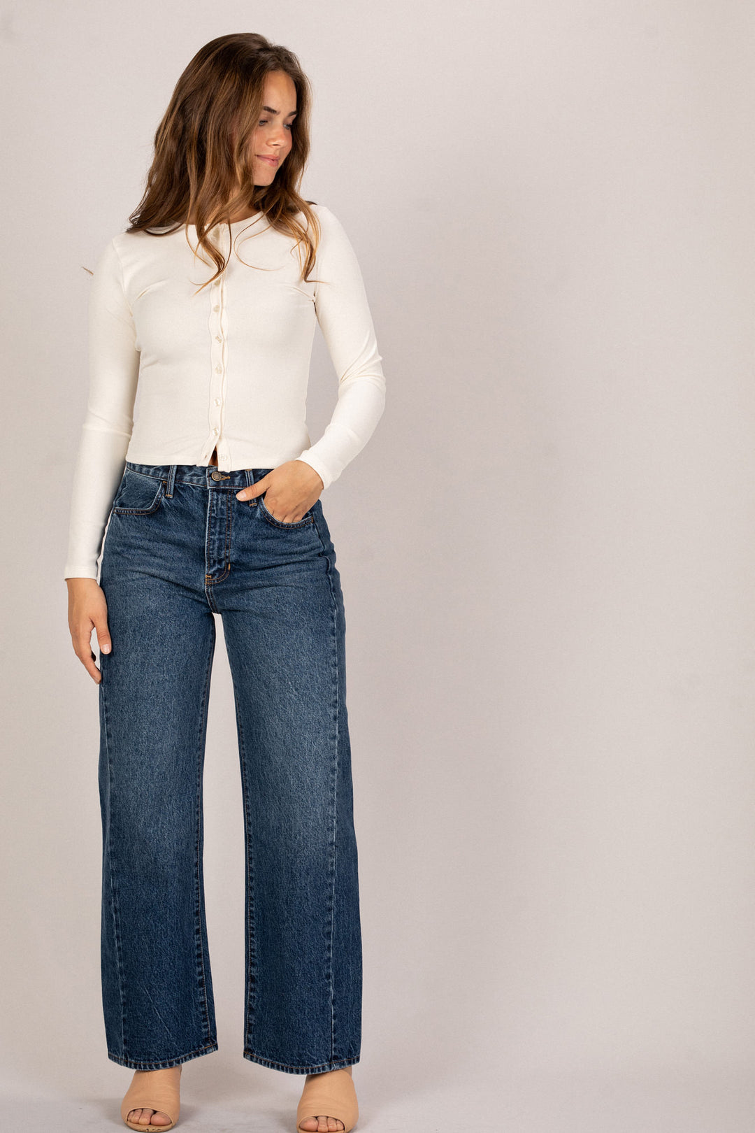 Woman wearing a white long-sleeved top, high-waisted blue denim jeans, and cream-colored sandals, standing against a plain background.