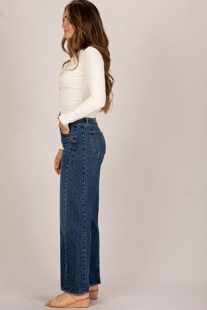 A young woman wearing high-waisted, straight-leg denim jeans in a dark wash, paired with a white fitted long-sleeve top and nude sandals.