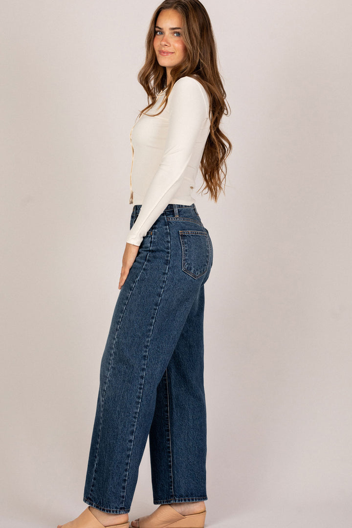 A young woman in a white top and high-waisted, straight-leg denim jeans, with long, wavy brown hair. The jeans have a barrel leg shape and a medium wash denim.