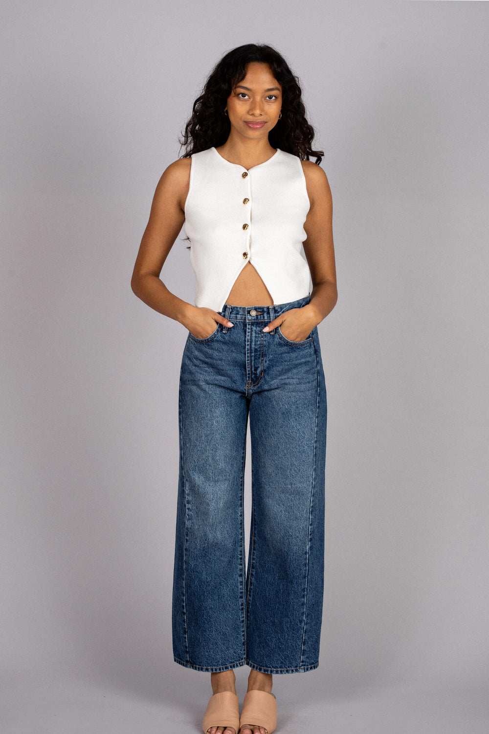 A young woman wearing a sleeveless white vest with brown buttons, paired with high-waisted blue jeans, posing against a gray background.