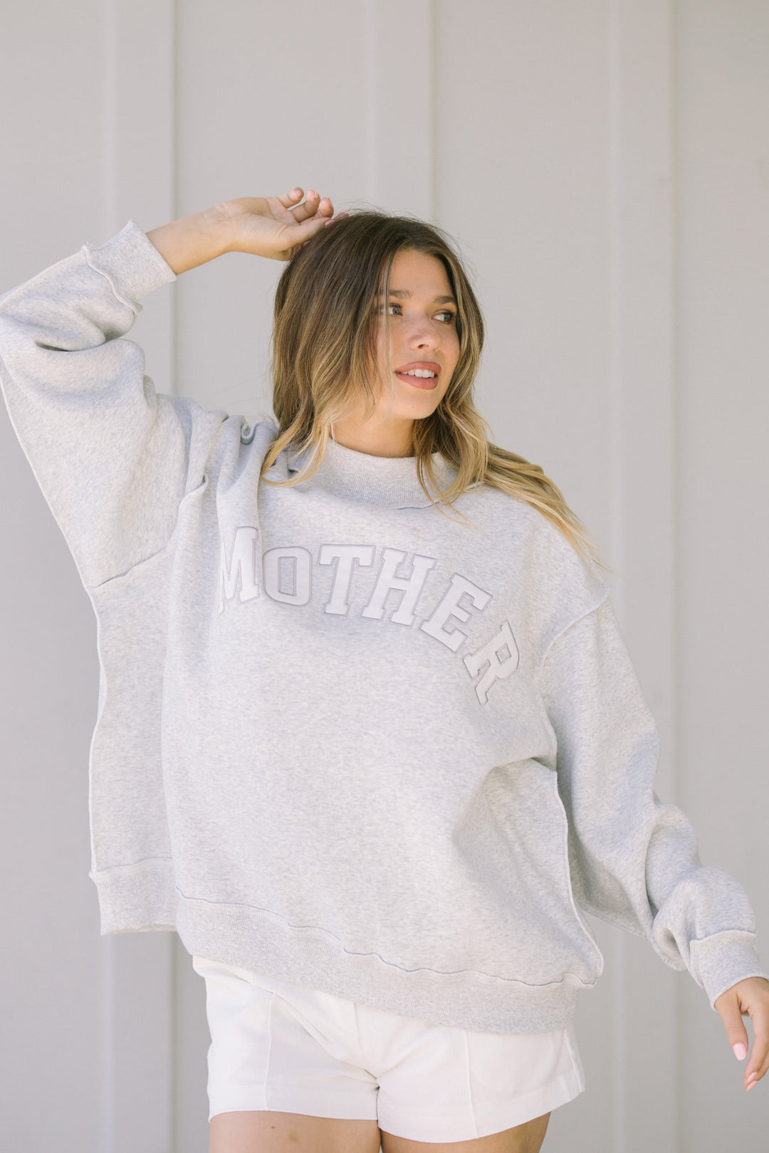 A grey, mock-neck sweatshirt with "MOTHER" printed in a stylish, distressed design. The model has blonde, wavy hair and is wearing the relaxed, cozy-looking top.