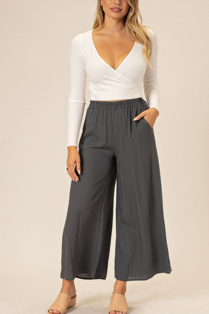 A young woman wearing a white long-sleeved top and gray wide-leg pants with pockets, standing against a neutral background.