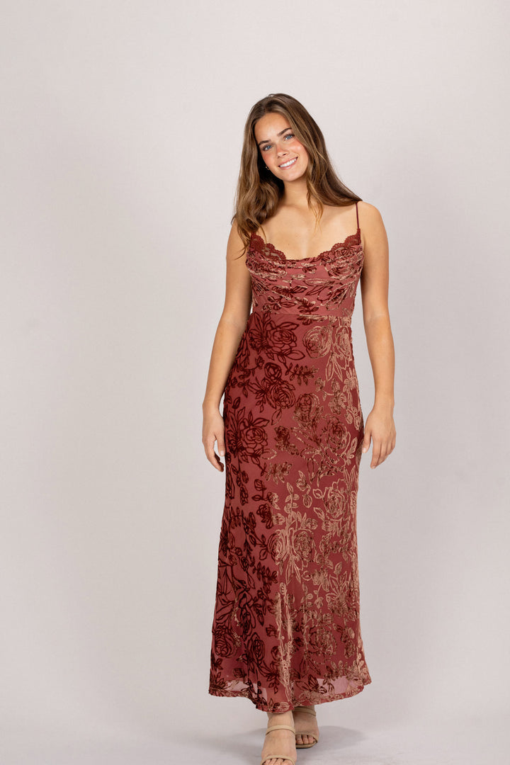A long, floral-patterned dress in a deep red color, with a cowl neckline and spaghetti straps. The dress has a fitted, flowing silhouette.