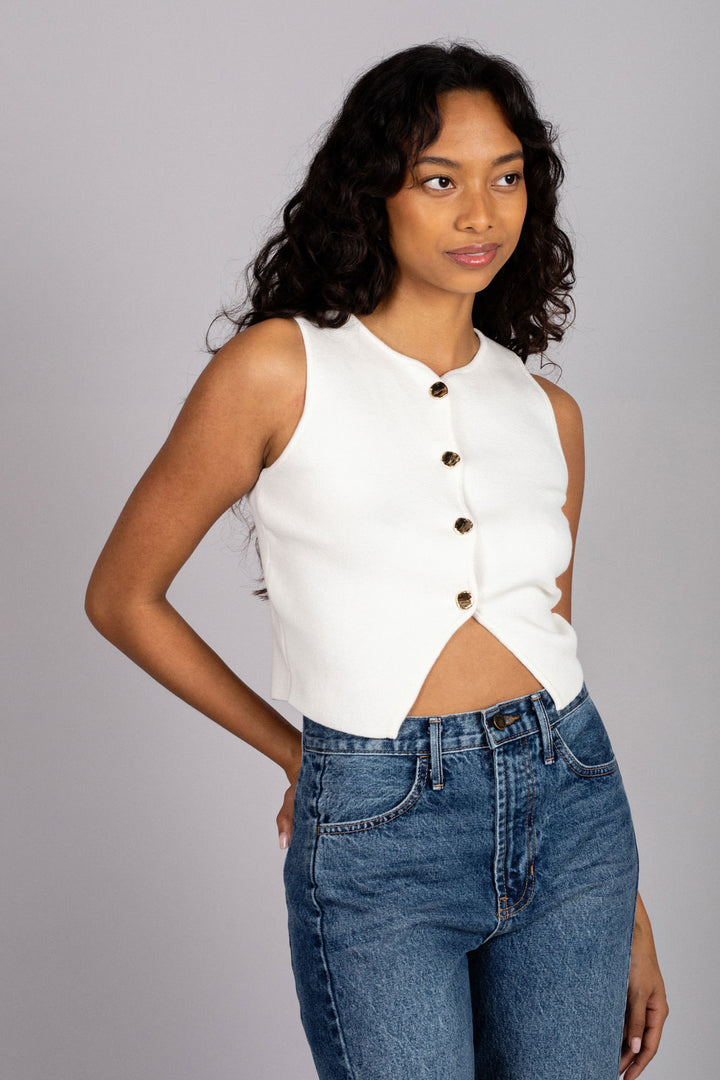 A sleeveless white buttoned vest top with a fitted silhouette, worn with high-waisted blue jeans by a woman with curly dark hair.