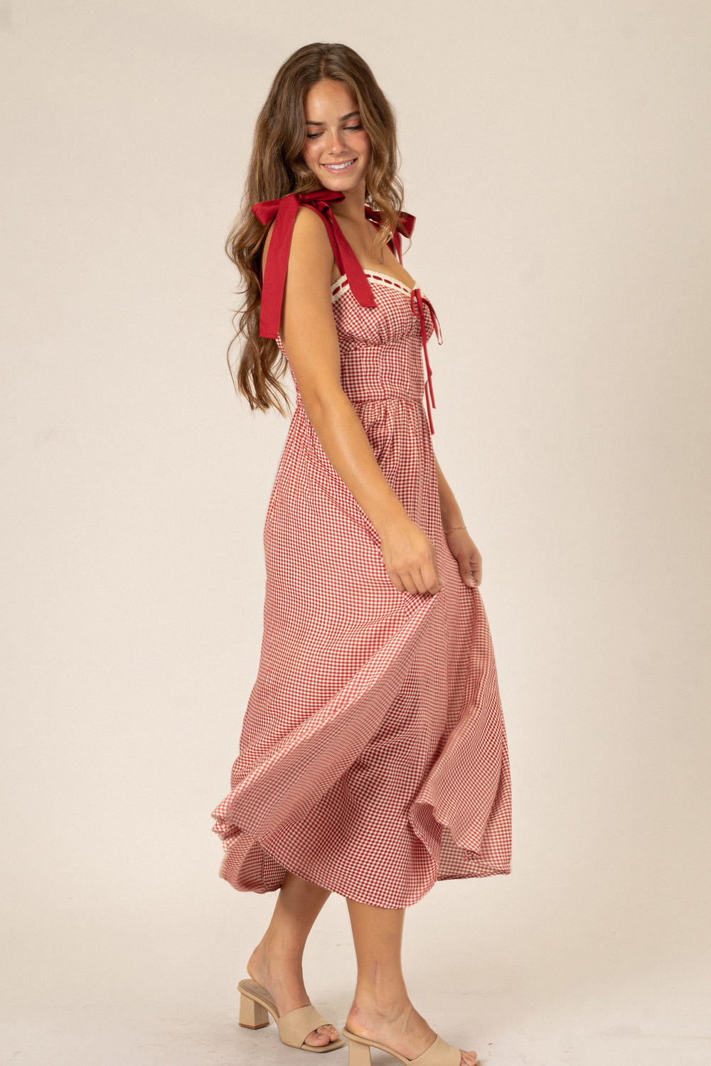 A red and white gingham print dress with a sweetheart neckline, ruched bodice, and midi-length skirt, modeled by a woman with long brunette hair.