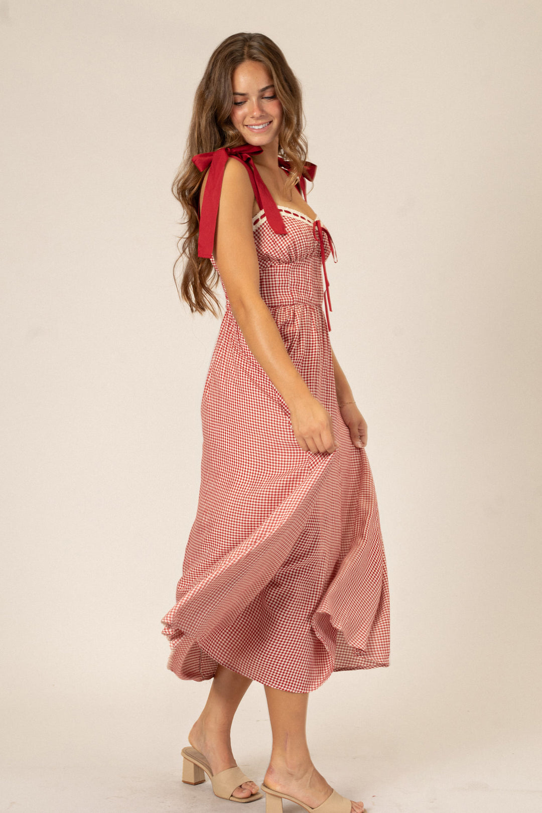 A red and white gingham print dress with a sweetheart neckline, ruched bodice, and midi-length skirt, modeled by a woman with long brunette hair.